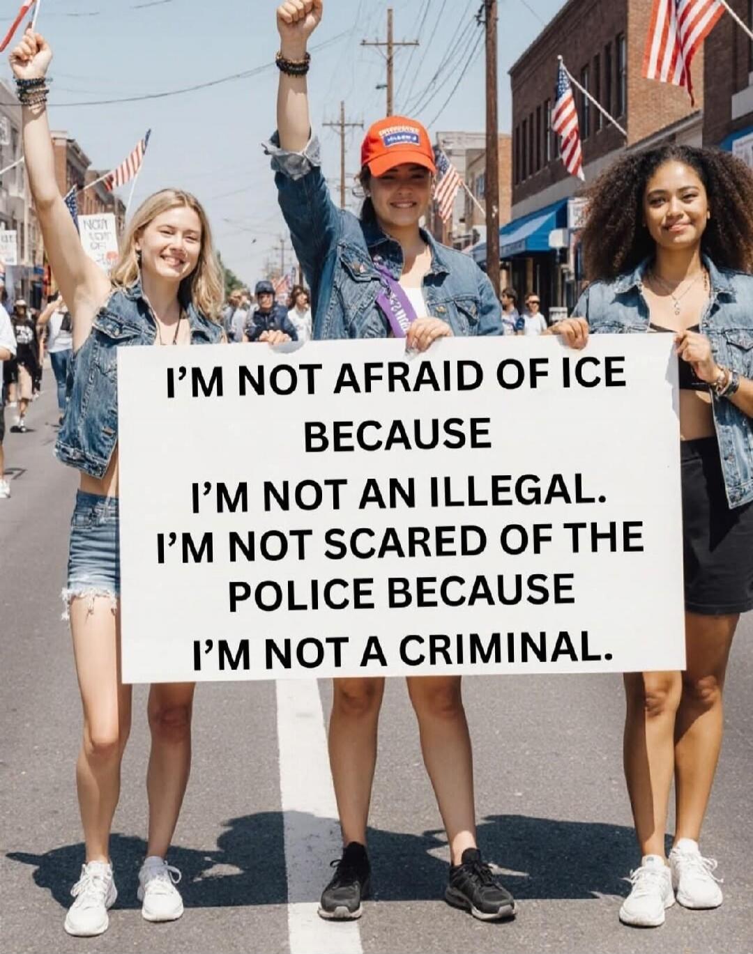 Three young women are holding a sign that reads: 'I'M NOT AFRAID OF ICE BECAUSE I'M NOT AN ILLEGAL. I'M NOT SCARED OF THE POLICE BECAUSE I'M NOT A CRIMINAL.' They are smiling and one has her fist raised in what appears to be a parade or protest with American flags in the background.