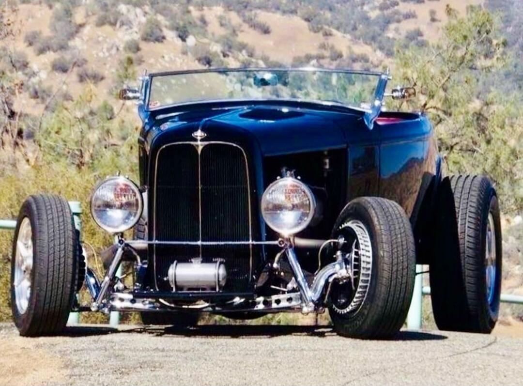 A classic black hot rod car with large tires and chrome details, parked outdoors on a sunny day with hills in the background.