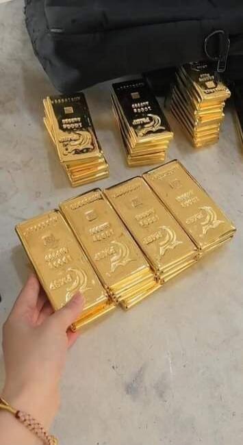 Stacks of golden bars are visible, some with text like 'GOLDEN BUDDHA' and '999.9 FINE GOLD'. A hand is holding one stack.