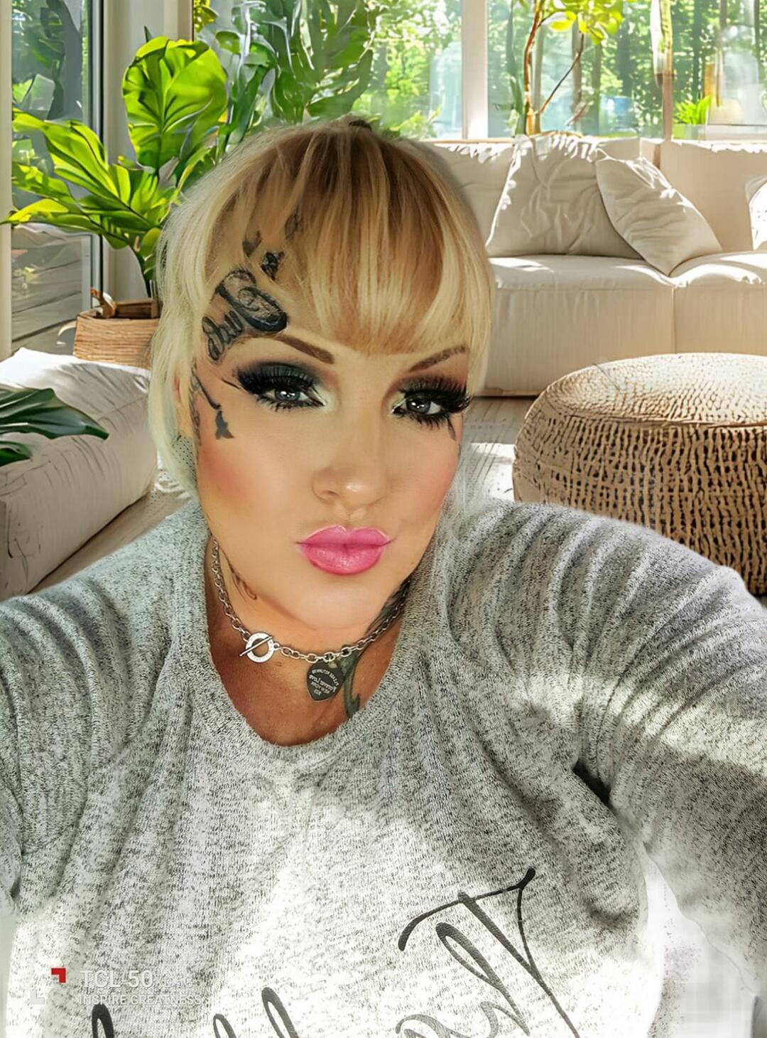 A person with dramatic makeup and tattoos posing for a selfie in a bright living room.
