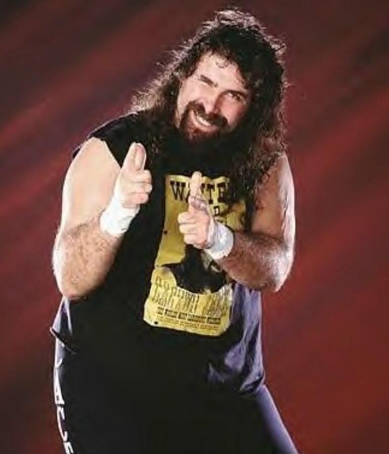 A muscular man with long dark hair and a beard, wearing a black sleeveless shirt with a yellow graphic, white wristbands, and pointing both index fingers toward the camera.