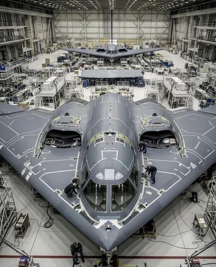 Two B-2 Spirit stealth bombers are seen in a hangar, with workers performing maintenance on one of them.