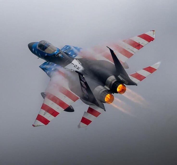 The image shows a fighter jet in flight, painted with the American flag motif. The jet is emitting smoke from its engines, suggesting it is performing a maneuver. The overall impression is one of power and patriotism.