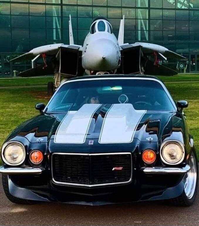 A classic black Chevrolet Camaro with white racing stripes parked in front of an F-14 Tomcat fighter jet on display.