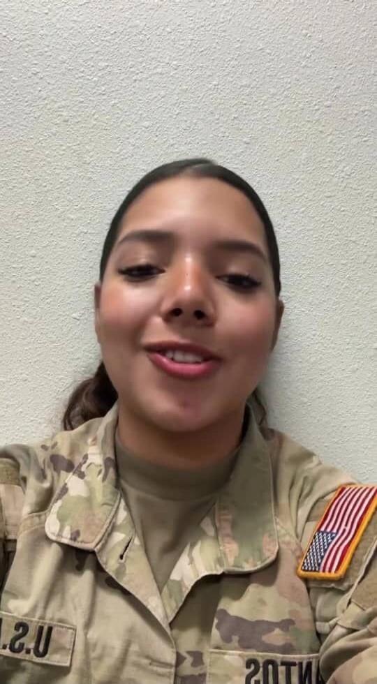 A woman in a US Army uniform is speaking directly to the camera, smiling. She appears to be sharing a message or an experience.