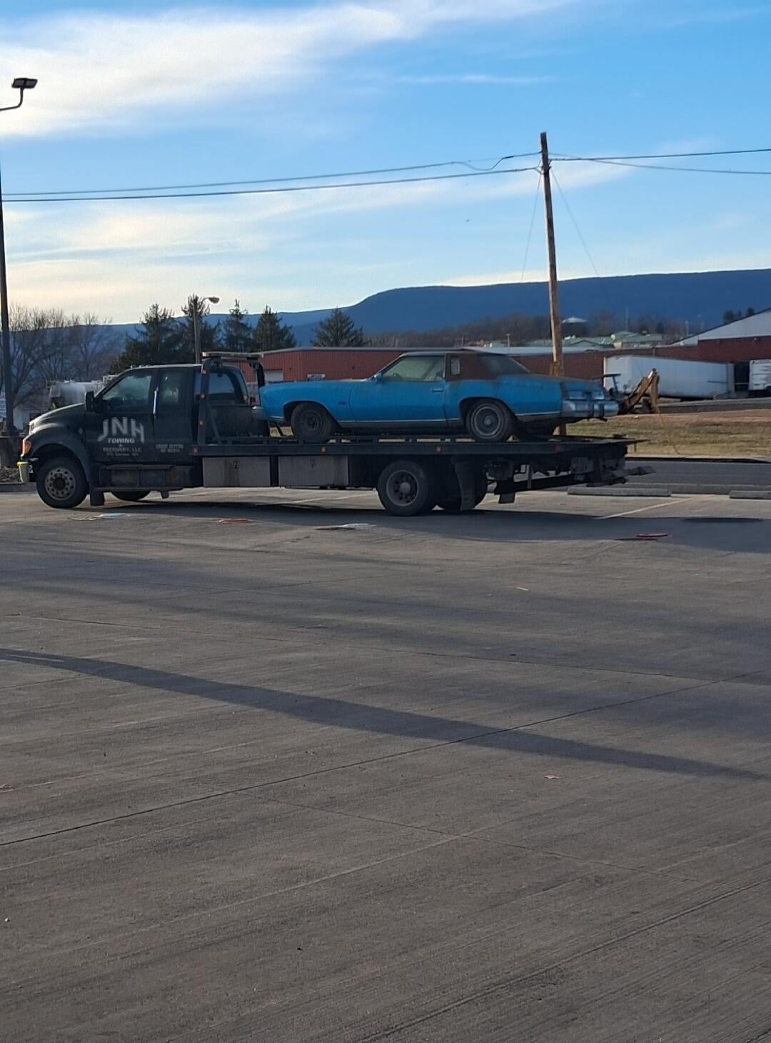 A tow truck hauls a blue vintage car on its flatbed along a paved road, with hills/mountains in the background.