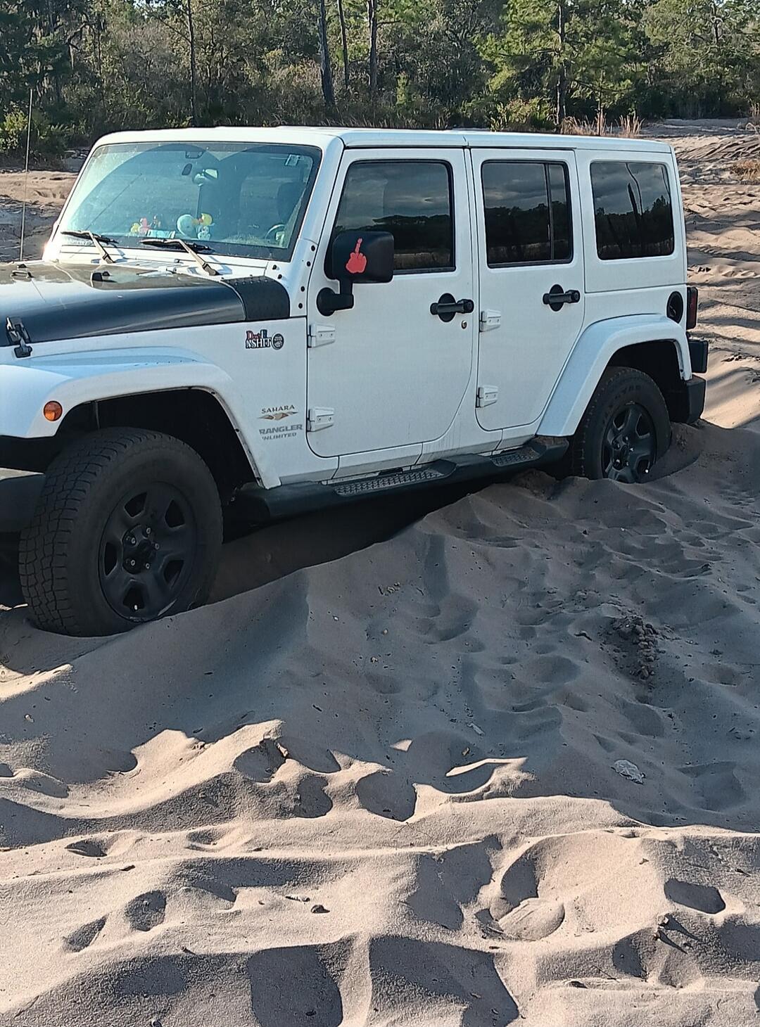 A white Jeep Wrangler Sahara Unlimited is stuck in deep sand. A small red middle finger emoji is visible on the side mirror.