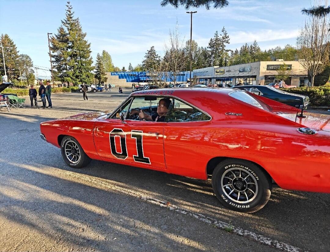 A bright orange classic Dodge Charger, resembling the General Lee from 'The Dukes of Hazzard' with '01' prominently displayed on its side, is driven by a person in a parking lot. The car features BFGoodrich Radial T/A tires and 'Charger' badging. In the background, other people and cars are visible, along with trees and buildings, including one wit