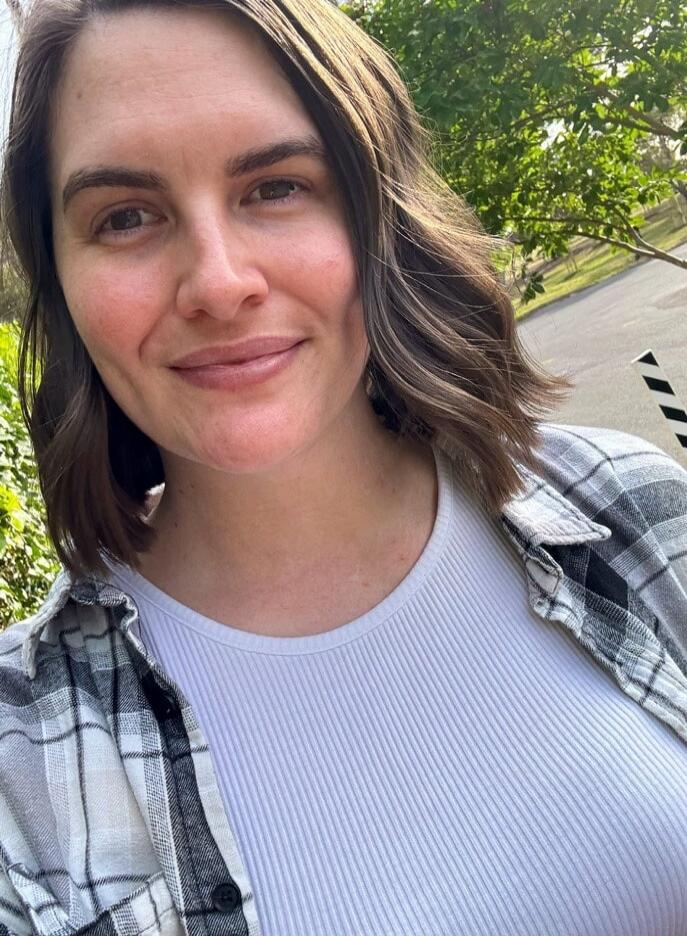A woman smiling at the camera, wearing a white ribbed top and a plaid shirt, outdoors with trees in the background.