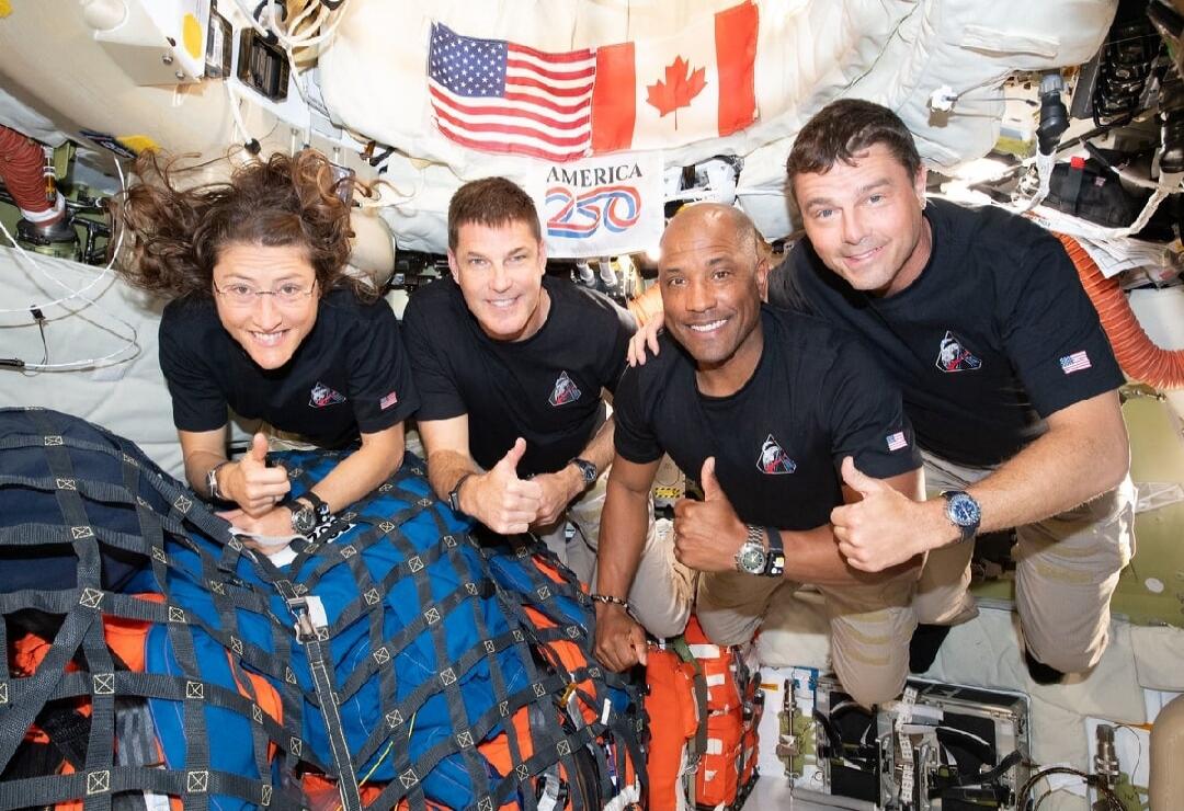 Four astronauts inside a spacecraft module, smiling and giving thumbs up. An American flag and a Canadian flag are visible in the background, along with the text 'AMERICA 250'.