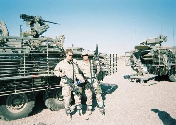 Two soldiers posing with their weapons in front of military vehicles.