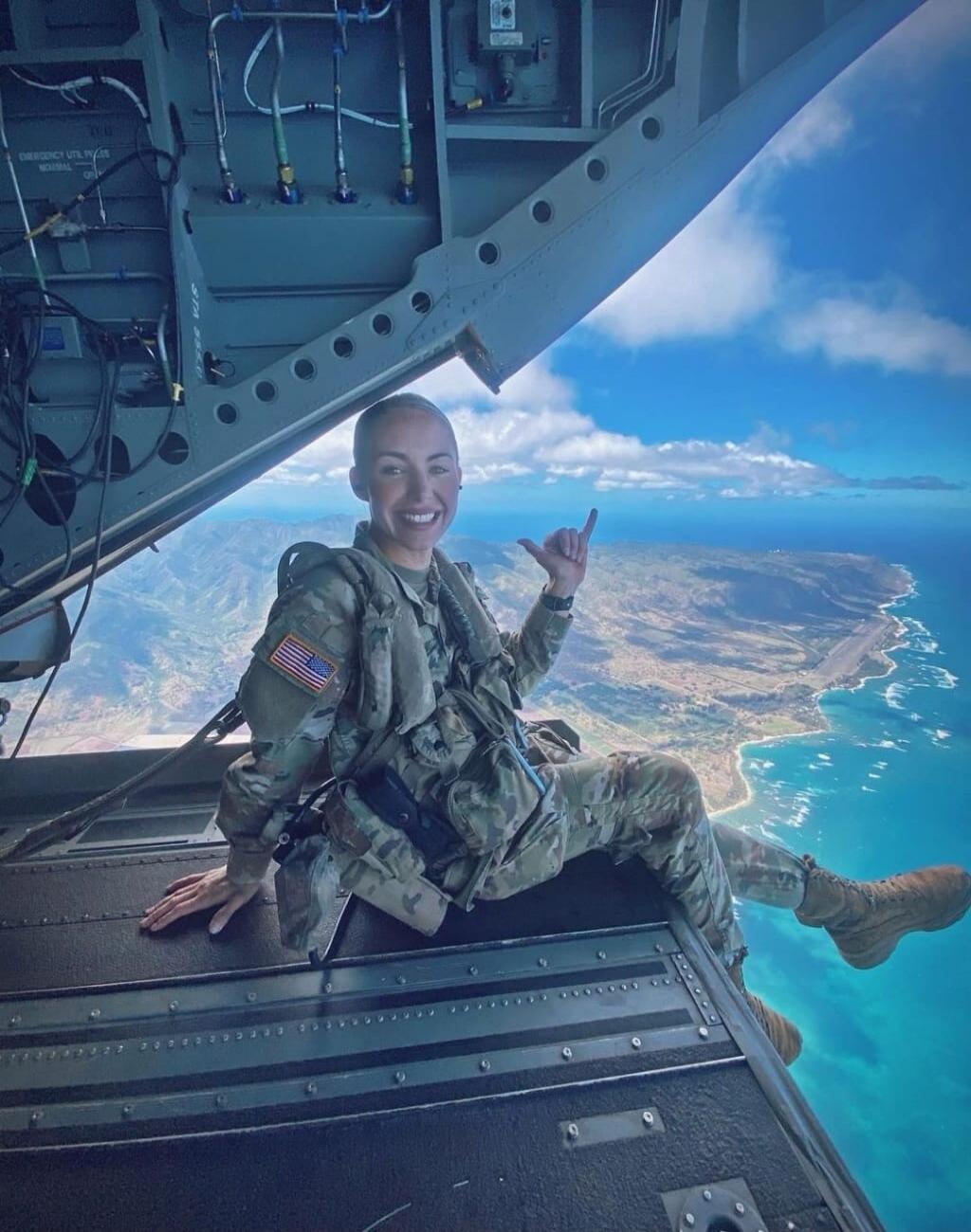 EMERGENCY UTL. P. GE NORMAL. A woman in military uniform is sitting on the edge of an open aircraft door, high above a beautiful coastal landscape with a clear blue ocean. She is smiling and making a 'shaka' hand gesture.