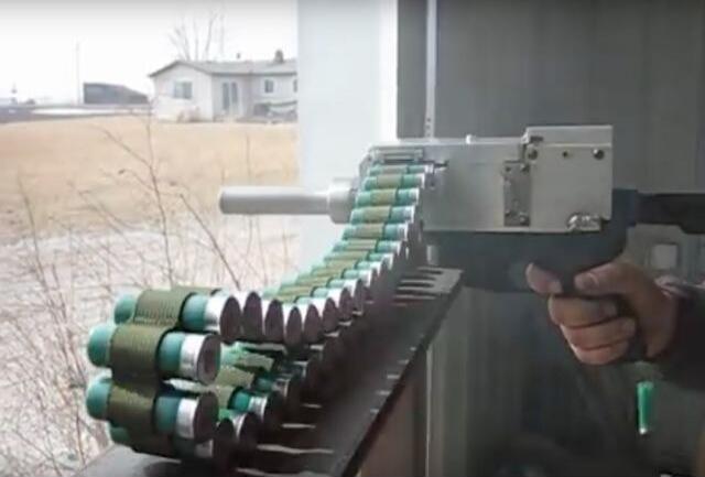 A person is holding a homemade belt-fed projectile launcher, possibly a potato gun, loaded with a long belt of small cans or tubes wrapped in green material. The device is aimed outdoors, with a house and bare trees in the background.