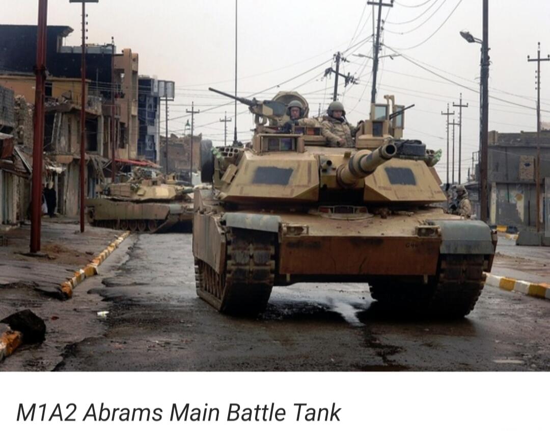 M1A2 Abrams Main Battle Tank. Two M1A2 Abrams tanks are seen on a damaged street in an urban area, with soldiers visible on the lead tank.