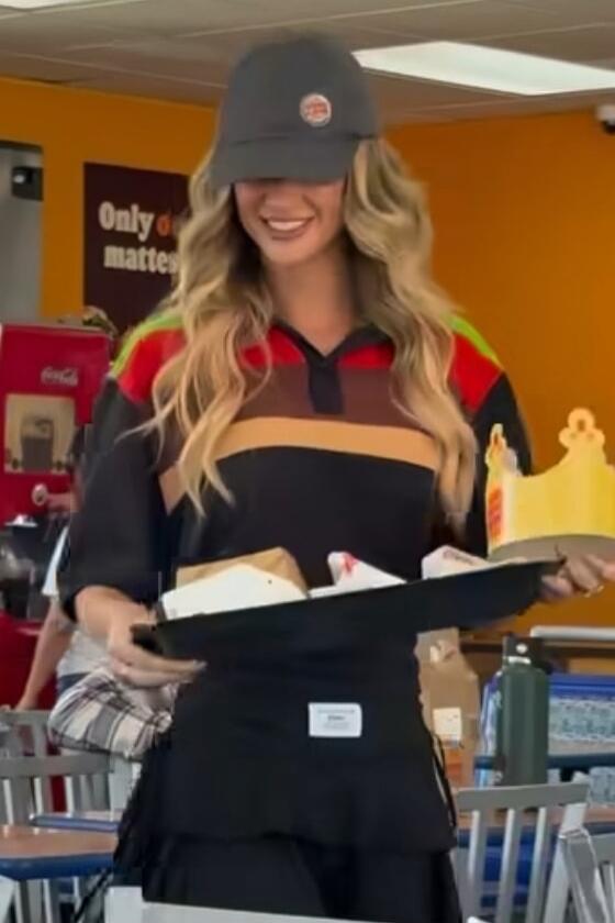 A young woman in a Burger King uniform, including a cap and a polo shirt with the brand's colors, is smiling while holding a tray with food items and a paper crown. A sign in the background partially reads 'Only matters'.