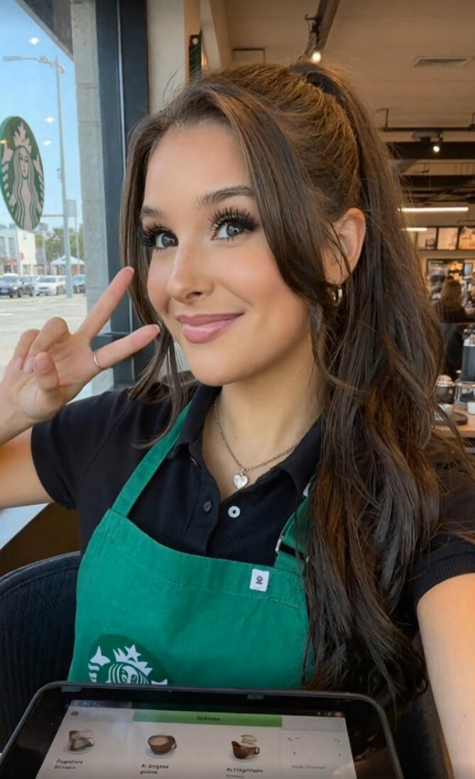 A young woman, likely a barista, smiling and making a peace sign while wearing a Starbucks apron and black polo shirt. She is inside a cafe, with a tablet showing menu items in front of her. A Starbucks logo is visible outside the window.