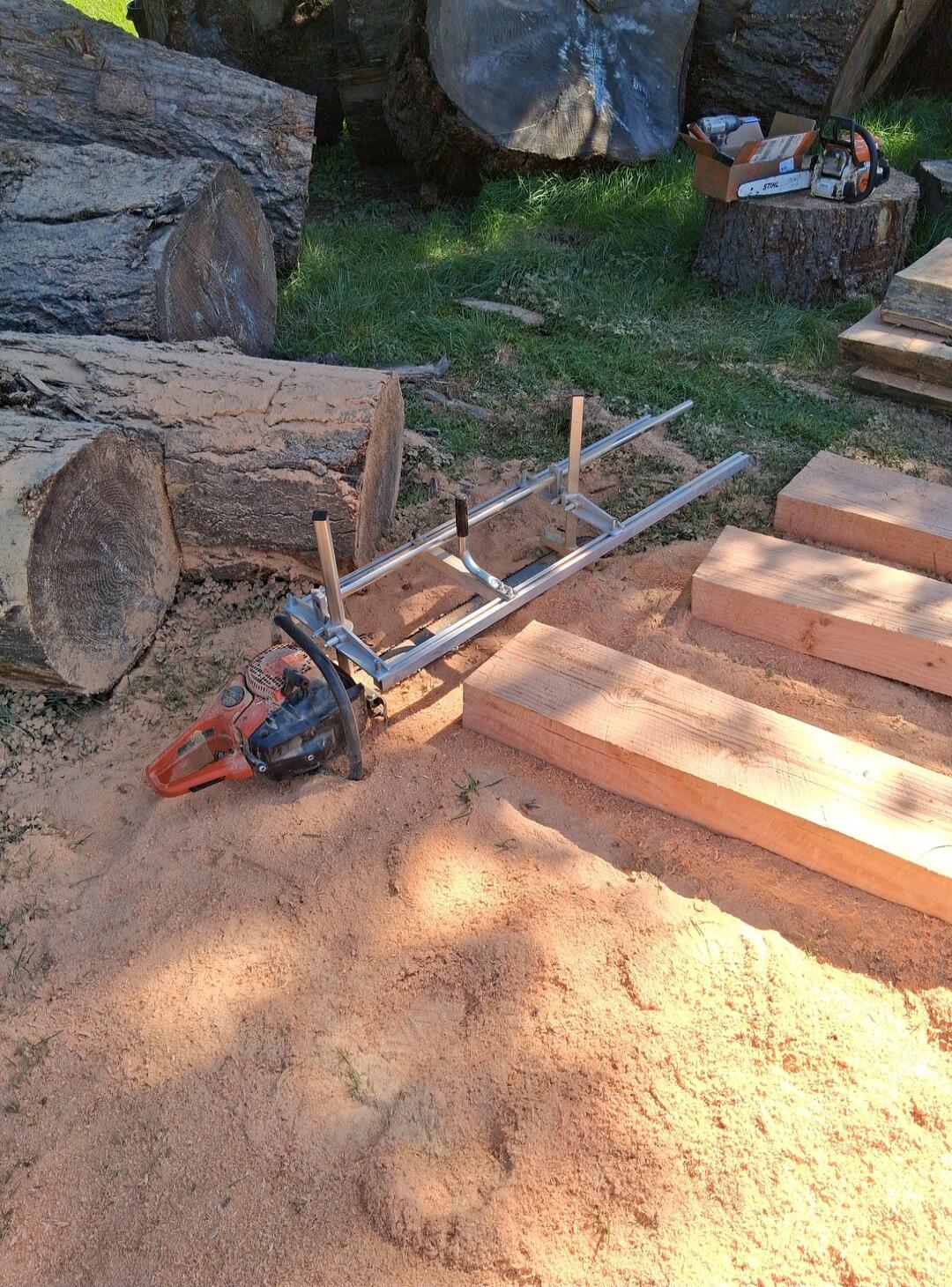 A chainsaw with a milling attachment is lying on a pile of sawdust next to large logs and freshly cut wooden boards. Another Stihl chainsaw and tools are visible in the background.