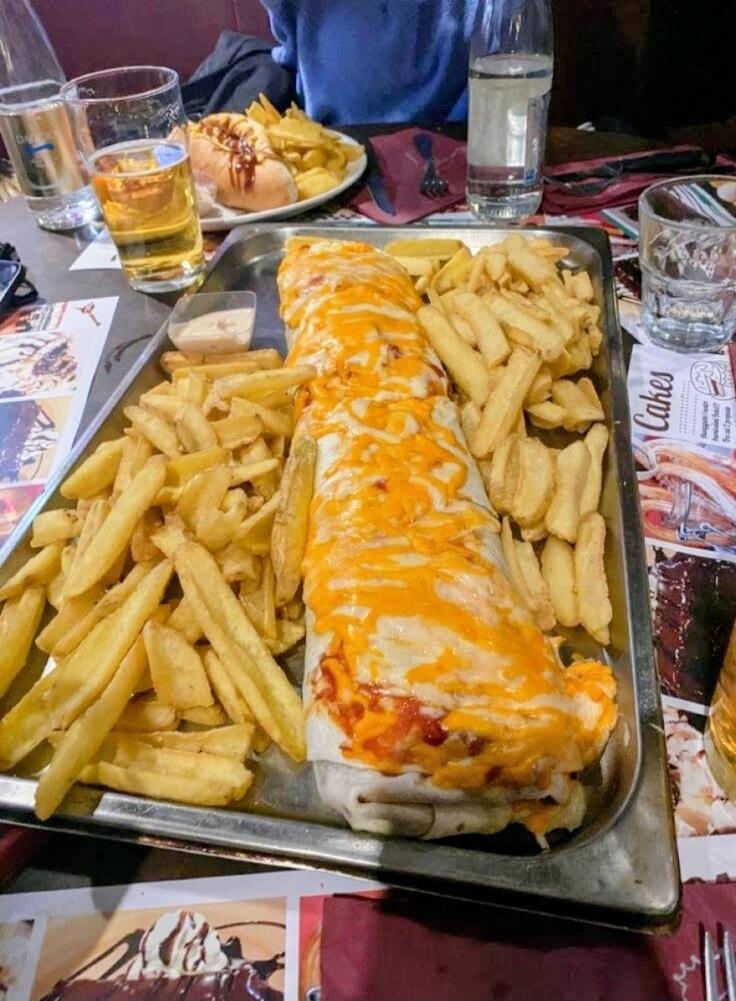 A large burrito topped with melted cheese, surrounded by french fries on a metal tray. In the background there are drinks and a plate with a sandwich. On a nearby surface, a magazine or menu shows the word 'Cakes' partially visible.