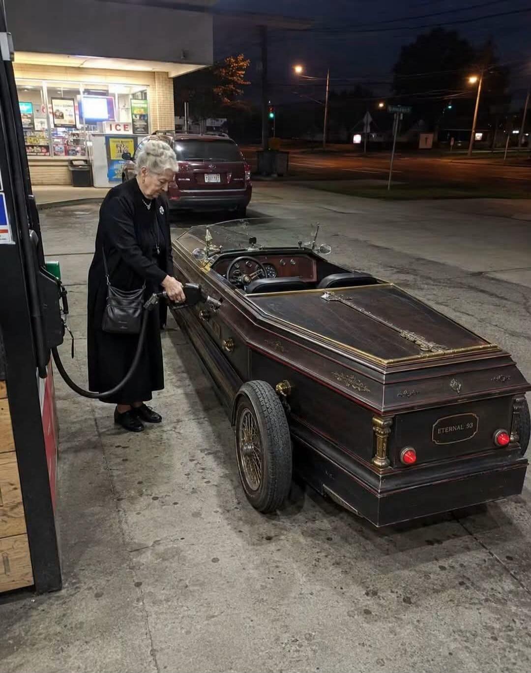ICE, ETERNAL 93. An elderly woman is fueling a custom car shaped like a coffin at a gas station.
