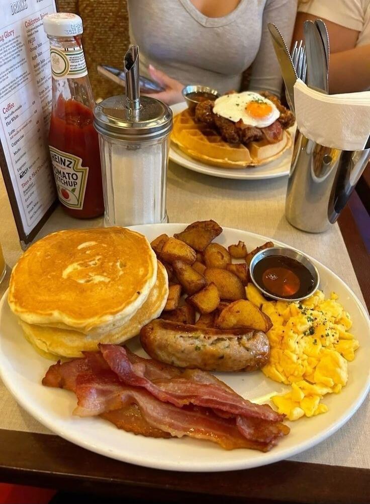 The image shows a delicious breakfast spread. In the foreground, a plate is filled with fluffy pancakes, crispy bacon, a juicy sausage, home fries, and scrambled eggs. A small dish of syrup or sauce is also visible. In the background, another plate features a waffle topped with fried chicken and a sunny-side-up egg. A bottle of Heinz ketchup and a 