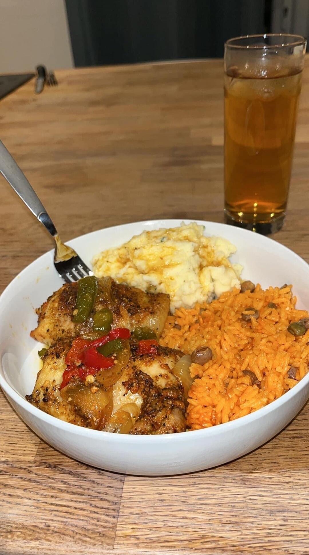 A close-up shot of a delicious homemade meal served in a white bowl on a wooden table. The meal consists of seasoned fish or chicken pieces topped with sautéed bell peppers and onions, a side of creamy mashed potatoes, and a portion of orange-colored rice with beans. A glass of iced tea is visible in the background.