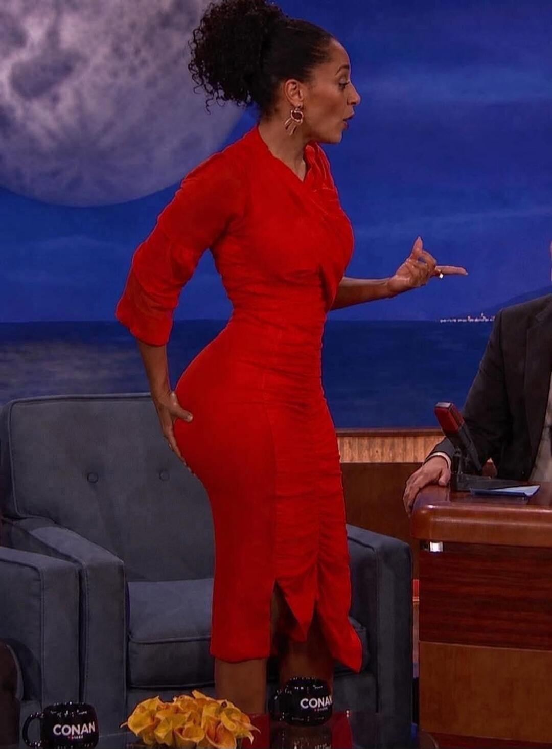 A woman in a red form-fitting dress on a talk show, playfully touching her backside and gesturing while speaking to the host.