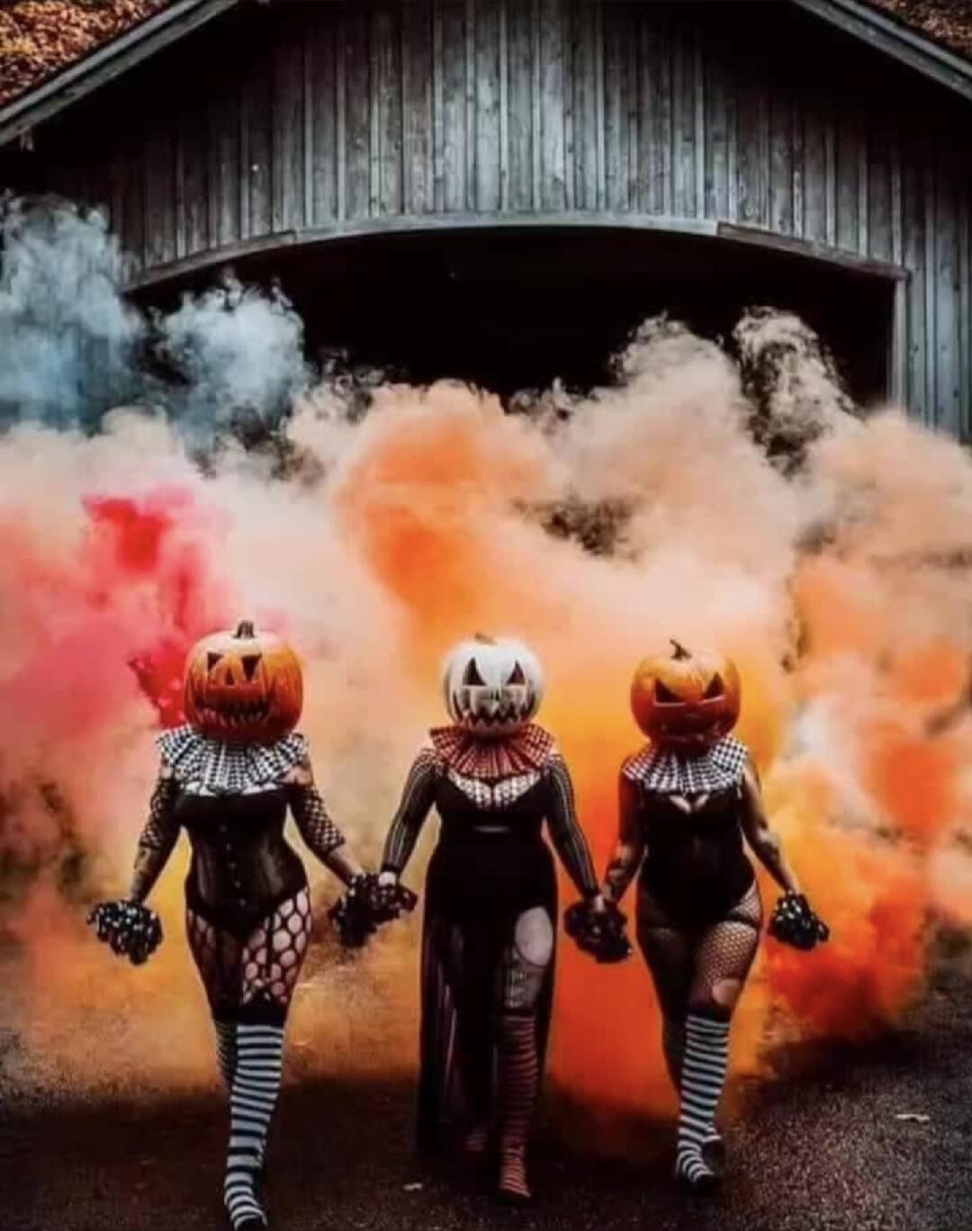 Three people in jack-o'-lantern heads wearing fishnet costumes walk hand in hand through colored smoke.