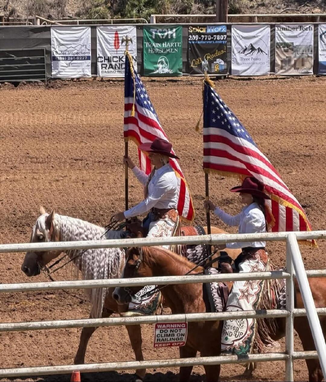 Two women on horseback, dressed in western attire including cowboy hats and chaps, are carrying American flags in a rodeo arena. In the background, there are various sponsor banners visible, including 'Bular Inspections (209) 268-9227', 'CHICKEN RANCH CASINO RESORT', 'TWAIN HARTE HORSEMEN', 'SolarWorx 209-770-7807 www.solarworx.energy', 'Let's Rode