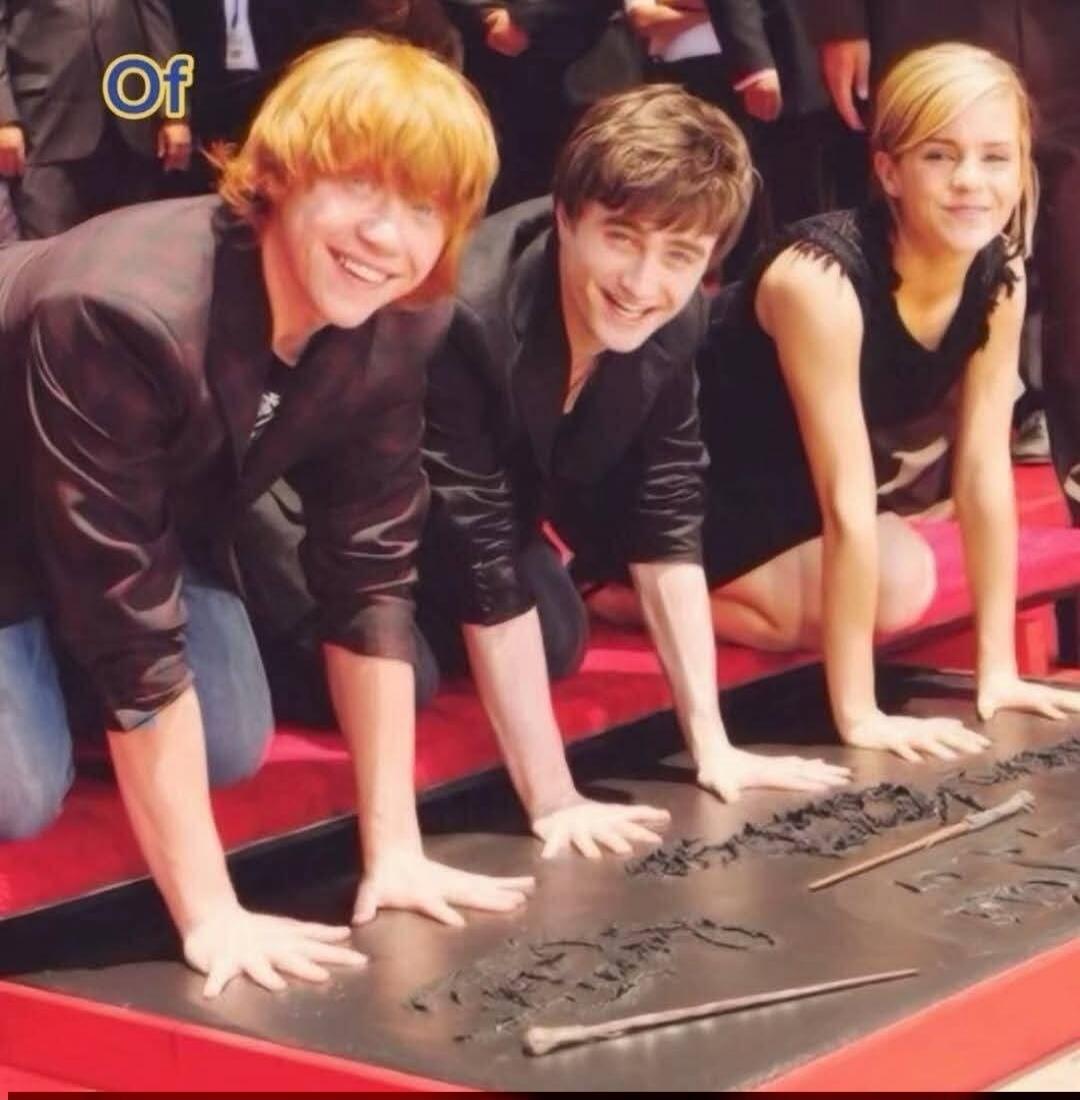 Of. The actors from Harry Potter, Rupert Grint, Daniel Radcliffe, and Emma Watson, are kneeling on a red carpet, smiling, and pressing their hands into a substance to create handprints. Two wands are also visible in the handprints.
