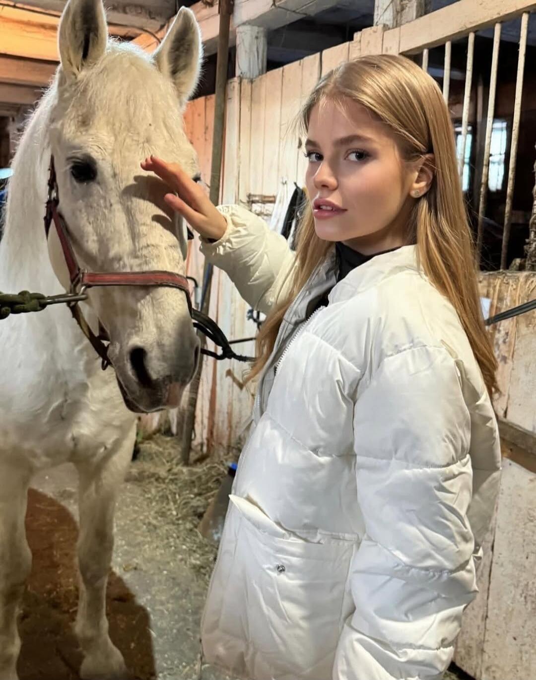A woman in a white puffer jacket touching a white horse in a stable.