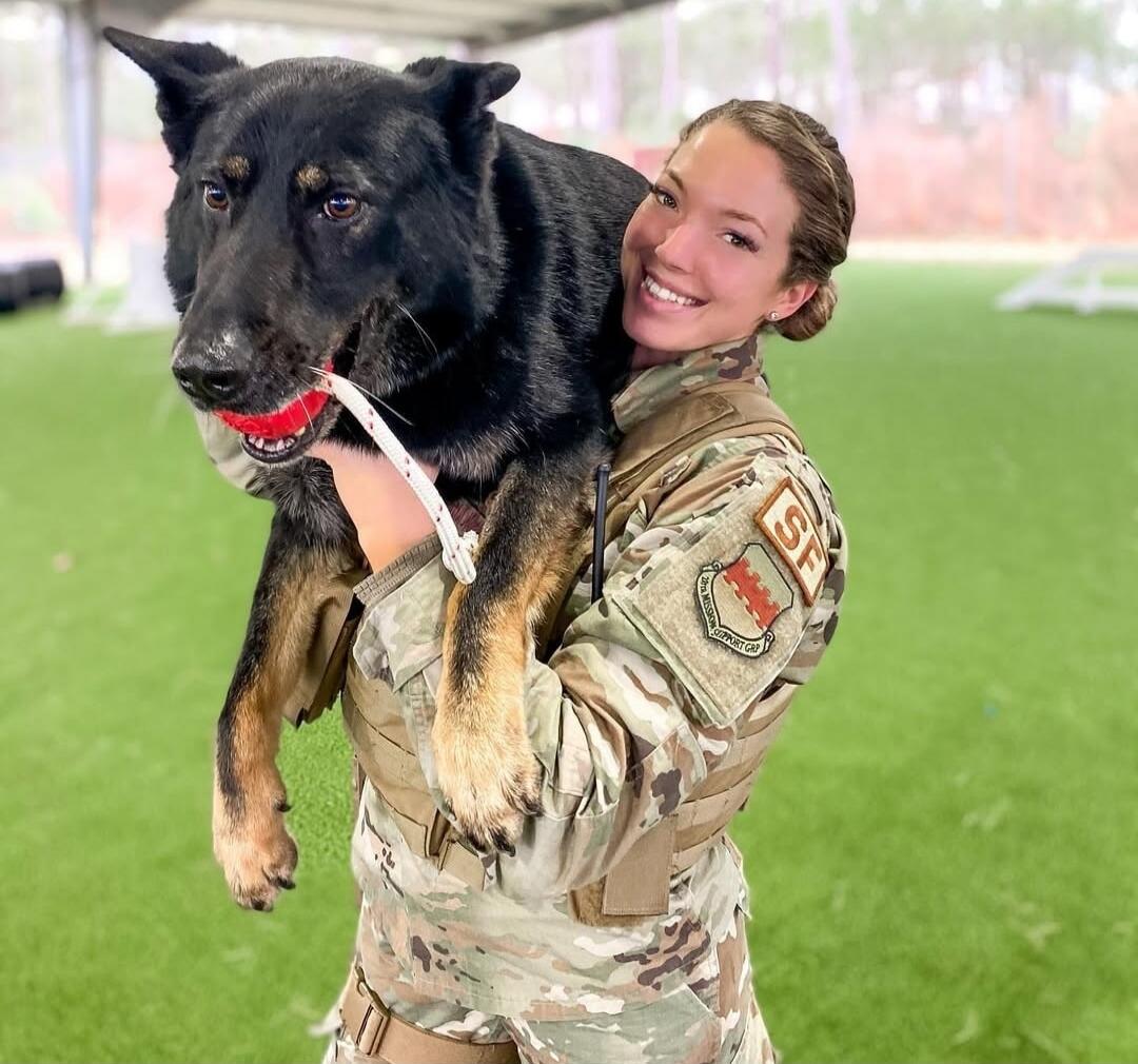 A smiling woman in a military uniform holds a large black and tan dog with a red ball in its mouth. The woman's uniform features a patch with 'SF' and '9TH MISSION SUPPORT GRP'.