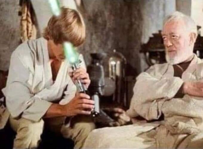 Luke Skywalker clumsily examining a green lightsaber, with the blade pointing towards his head, while Obi-Wan Kenobi watches him with a concerned expression.