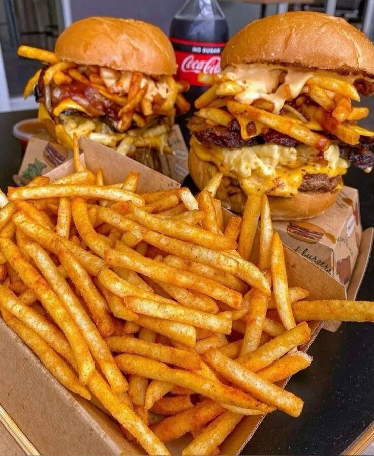 Two loaded burgers with melted cheese and toppings, served with a large pile of seasoned french fries. A bottle of Coca-Cola is in the background.