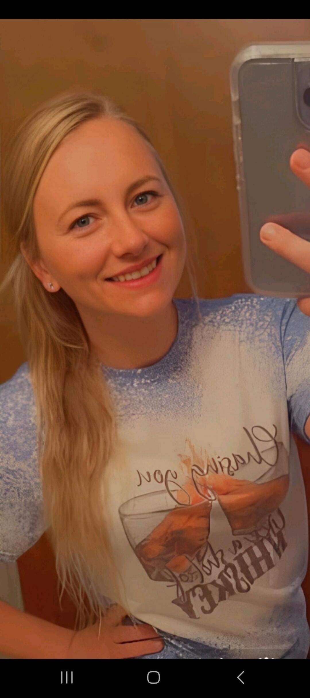 A blonde woman smiling and taking a selfie in a mirror. She is wearing a t-shirt with a graphic design that includes the word 'WHISKEY' and what appears to be 'pour' or similar text, along with an image of a drink being poured into a glass.