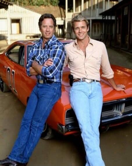 Two men, likely actors from 'The Dukes of Hazzard', posing with the iconic orange car 'General Lee'. One man wears a blue plaid shirt and jeans, the other a light-colored shirt and light blue jeans. The number '01' is visible on the car door.
