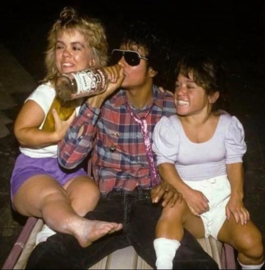 Michael Jackson (or look-alike) drinking from a Jack Daniel's bottle, flanked by two women with dwarfism. The bottle clearly shows 'JACK DANIEL'S OLD No. 7 BRAND'.