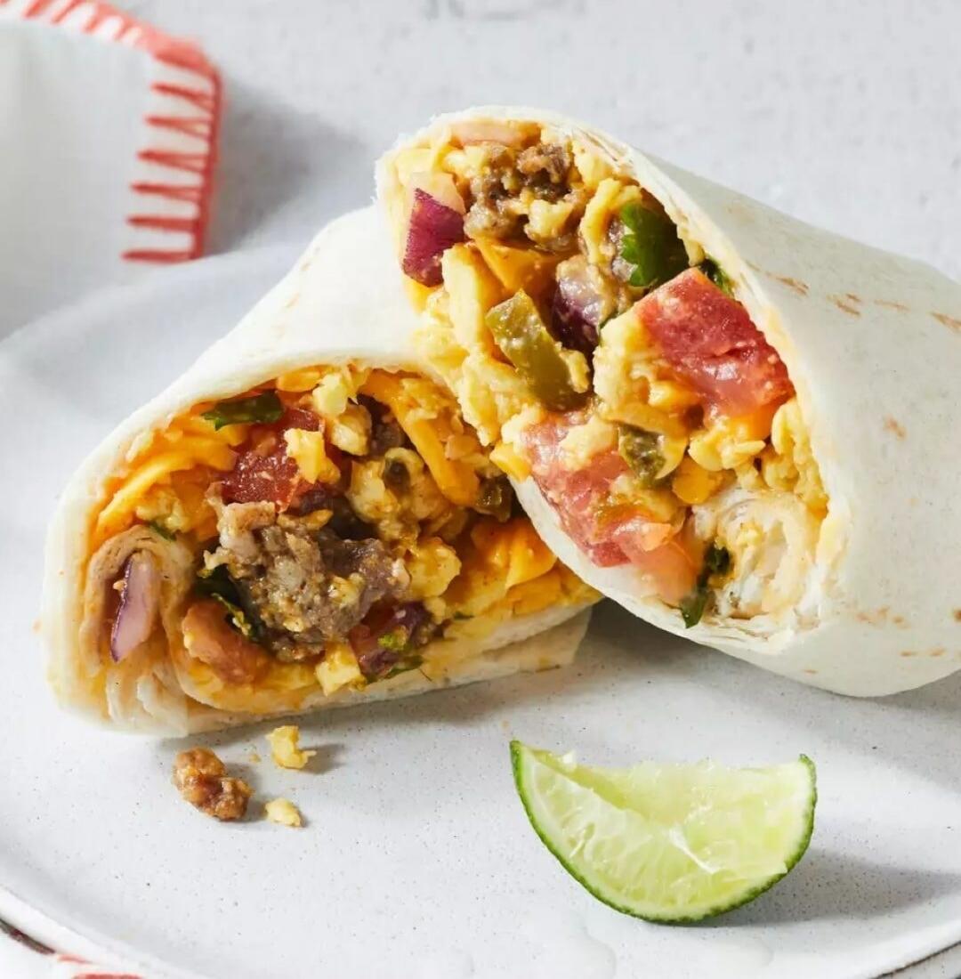 A breakfast burrito cut in half, revealing its rich filling of scrambled eggs, cheese, sausage or ground meat, diced tomatoes, red onions, and green peppers. A lime wedge is placed next to it on a white plate.
