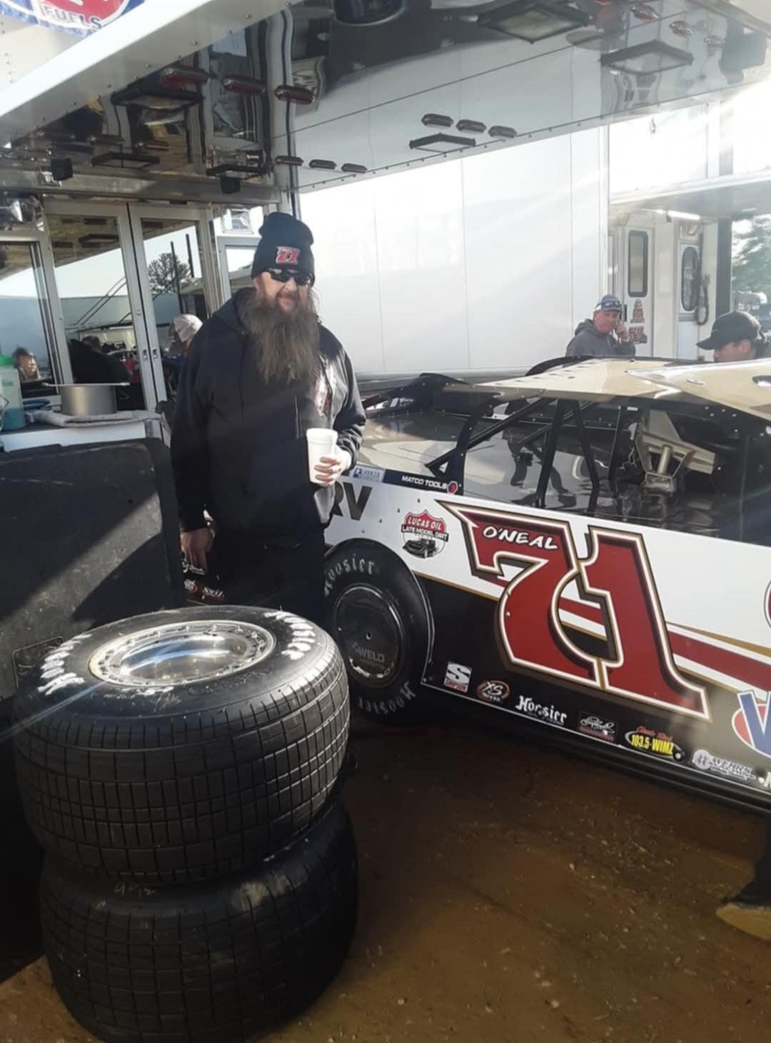 A man with a long beard and sunglasses, wearing a black hoodie and a beanie with '71', holds a cup. He stands next to a black and white dirt track late model race car, also numbered '71', featuring sponsors like 'O'NEAL', 'LUCAS OIL LATE MODEL DIRT', 'MATCO TOOLS', 'Hoosier', '103.5 WIMZ', 'S', and 'XS'. Two Hoosier racing tires are stacked in the 