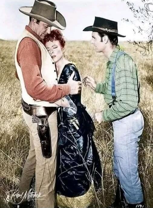 A vintage Western scene with a man in a cowboy hat holding a woman as another man in a hat stands nearby in a field.