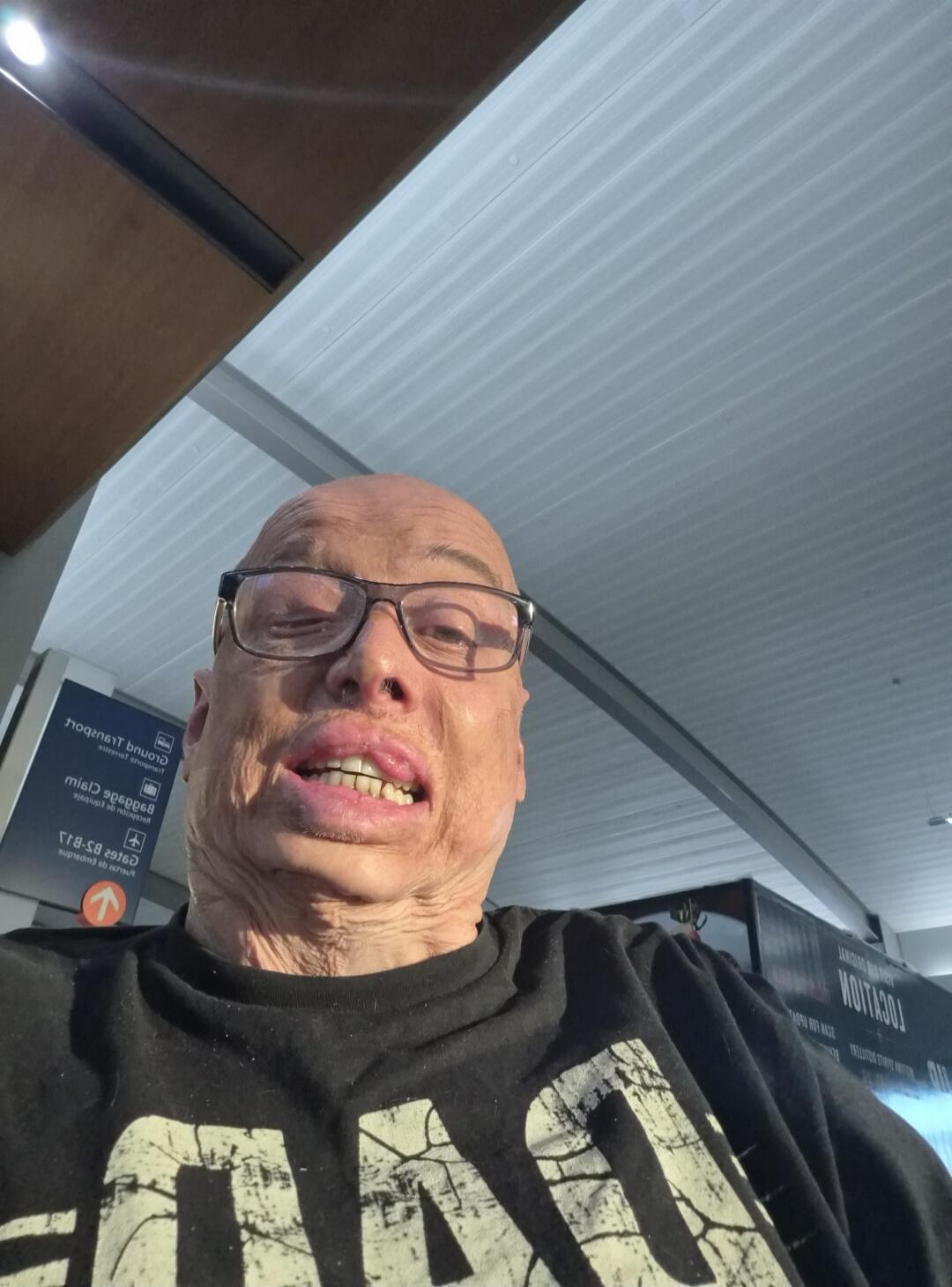 A person with a distinctive facial appearance, wearing glasses and a black t-shirt with 'DAD' written on it, making an unusual facial expression. In the background, there are signs that appear to be in an airport or public transport hub, with words like 'TRANSPORT', 'LOCATION', and 'Claim baggage' visible.