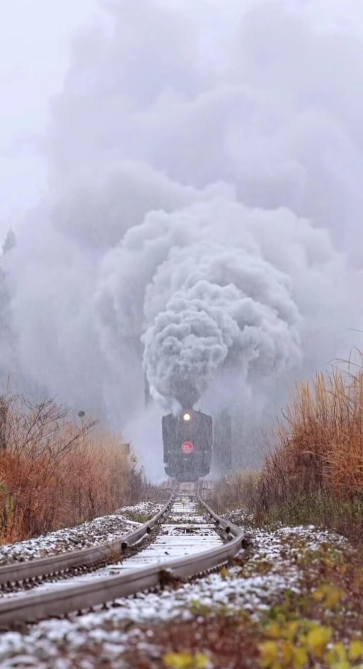 A steam locomotive on a railway track releasing a large plume of smoke, in a snow-dusted rural setting.