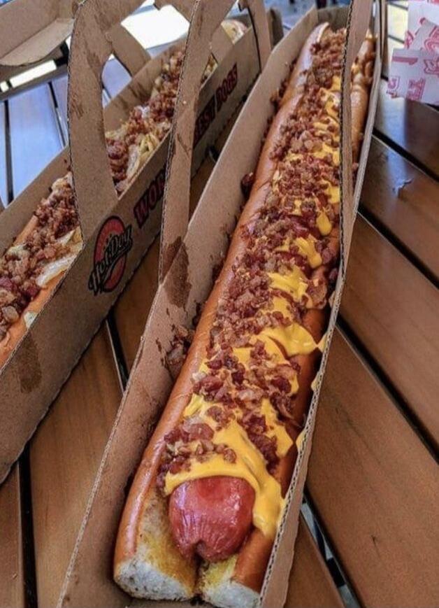 Two oversized hot dogs topped with melted cheese and crumbled bacon, served in cardboard takeout boxes.