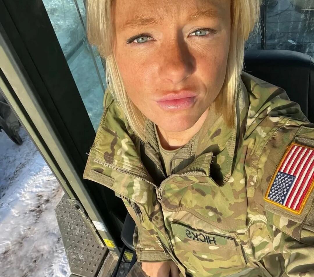 A blonde woman in a military uniform, with an American flag patch on her sleeve, takes a selfie inside a vehicle with snow visible outside. Her name tag reads 'HICKS'.