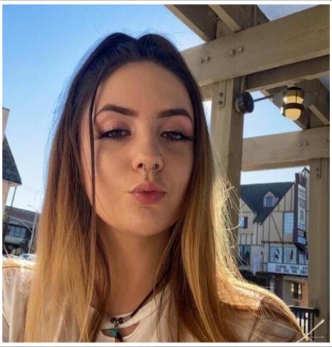 A young woman takes a selfie outdoors, with long hair and light makeup, standing under a wooden structure.