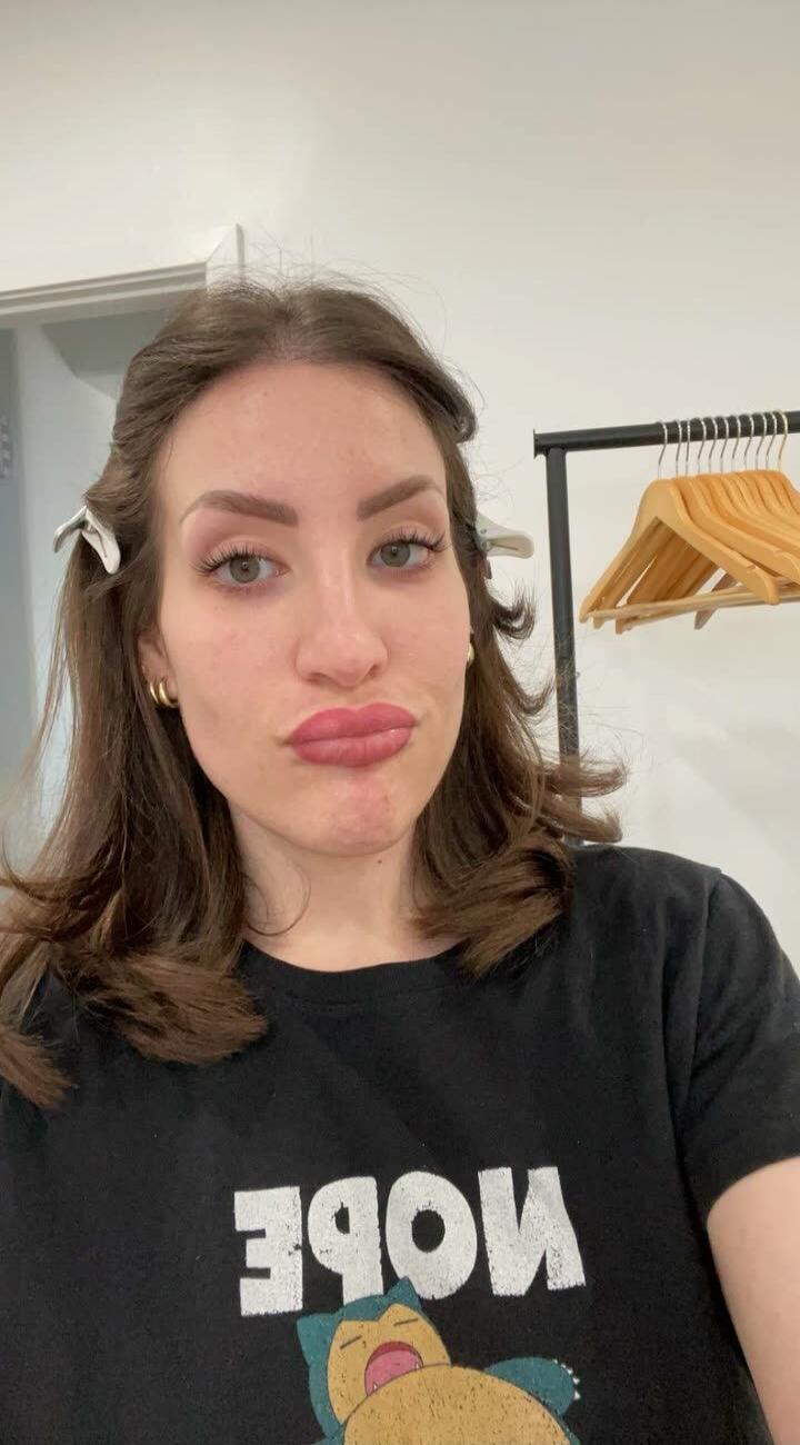 A woman in a black t-shirt with a Snorlax print and the word 'MODE' (reversed) on it, with hair clips in her hair, pouting for a selfie.
