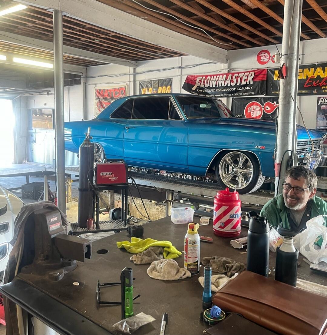 Flowmaster banners on wall; blue vintage car on a lift in a workshop; tools and bottles scattered on the workbench; a man is smiling nearby.