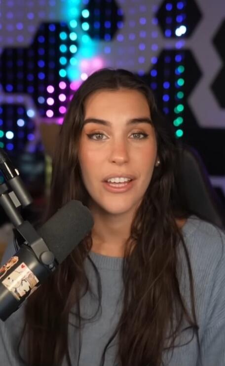 A young woman with long dark hair is speaking into a microphone in a room with decorative LED lights in the background. She appears to be a streamer or podcaster.