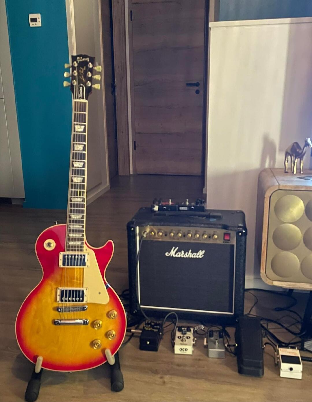 Sunburst Les Paul-style electric guitar on a stand, connected to a Marshall amplifier with several guitar pedals on the floor. A blue wall and a cabinet with tuning pegs visible in the background.