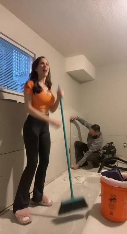 A woman in an orange crop top is sweeping a dusty room while a man works on the wall. She appears to be singing or talking, possibly about the ongoing home renovation or the task of cleaning.