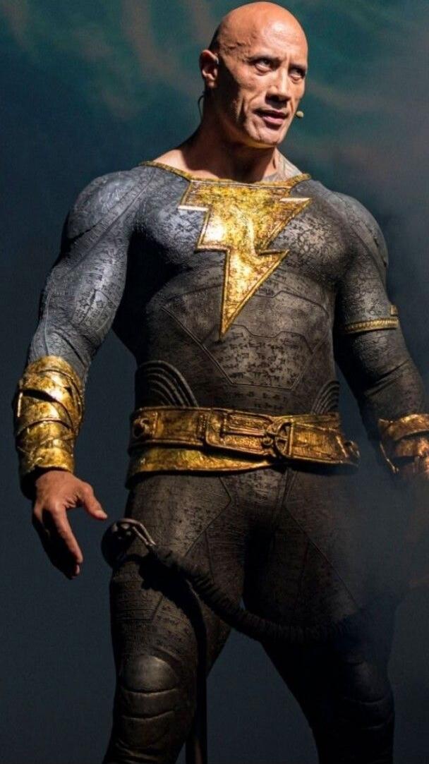 A man in a dark, textured superhero suit with a prominent gold lightning bolt emblem on the chest, gold wristbands, and a gold belt. He stands looking ahead.